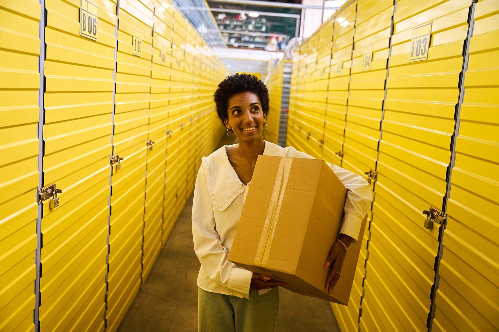 Woman with short curly hair carrying a cardboard box in a warehouse corridor lined with yellow storage lockers.